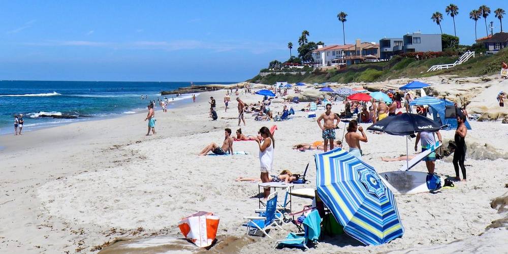 A busy San Diego beach on a clear day, with umbrellas, surfboards and houses along the bluff above.